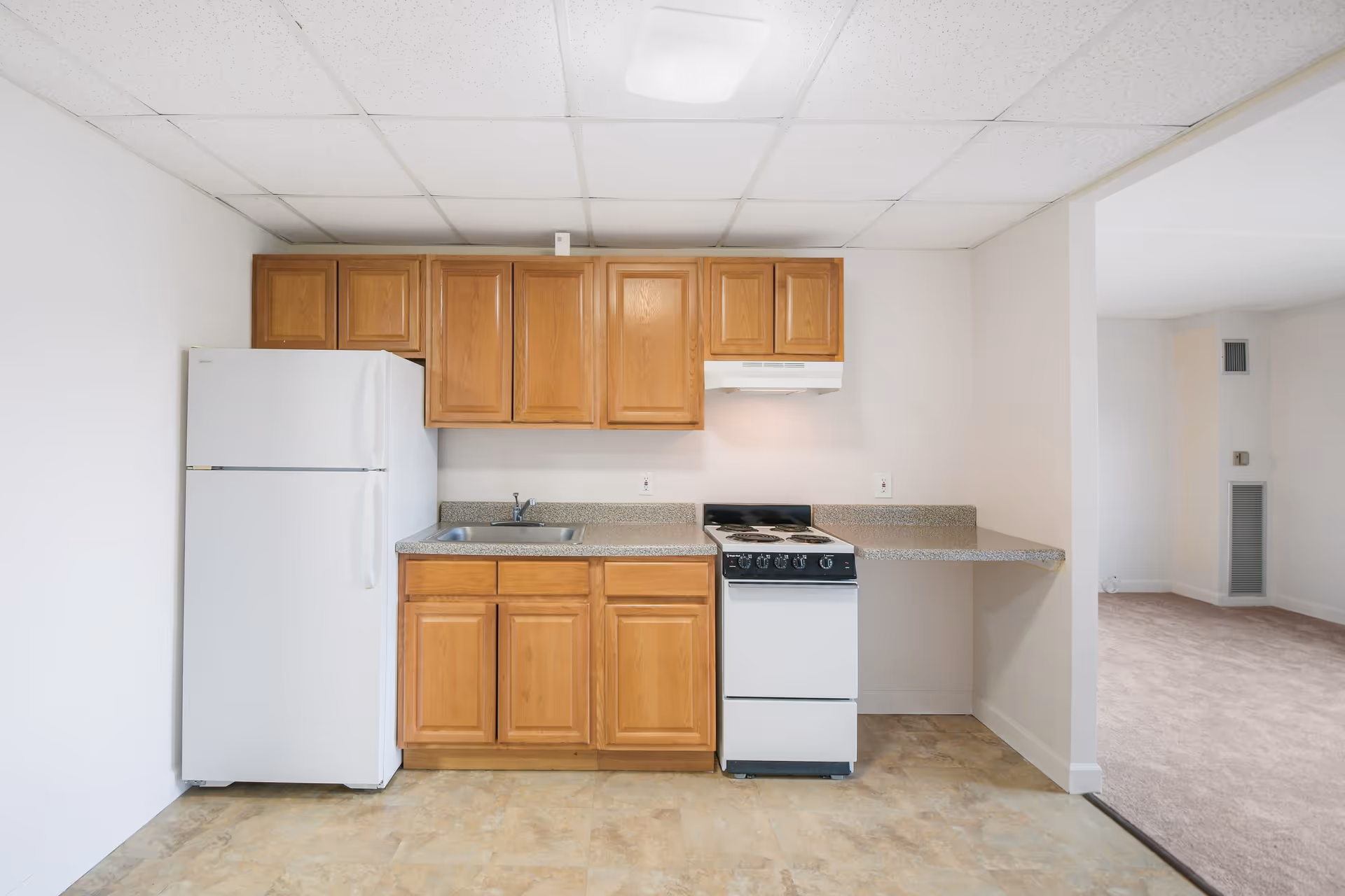 A small kitchen area with wooden cabinets, a white refrigerator, a stainless steel sink, a white stove with an overhead vent, and a countertop extending to the right. The kitchen floor has a beige tile pattern, and the adjacent room has carpeted flooring.