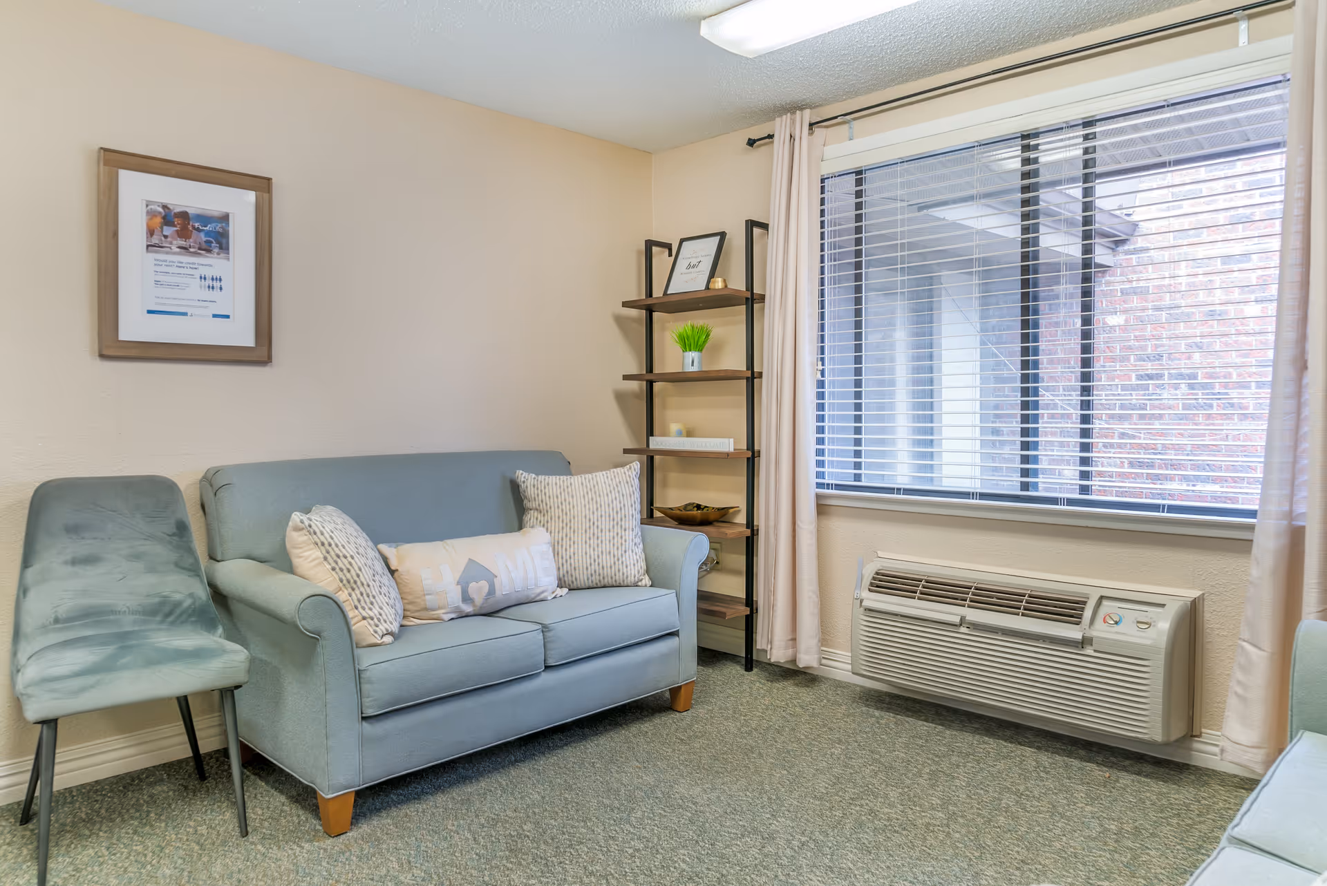 Small sitting area with a light blue loveseat, side chair, shelving unit, and a window air conditioner beneath a large window.