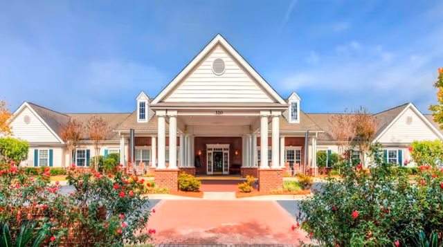 Front entrance of a single-story senior living building with a columned portico, gabled roof, and landscaped driveway.