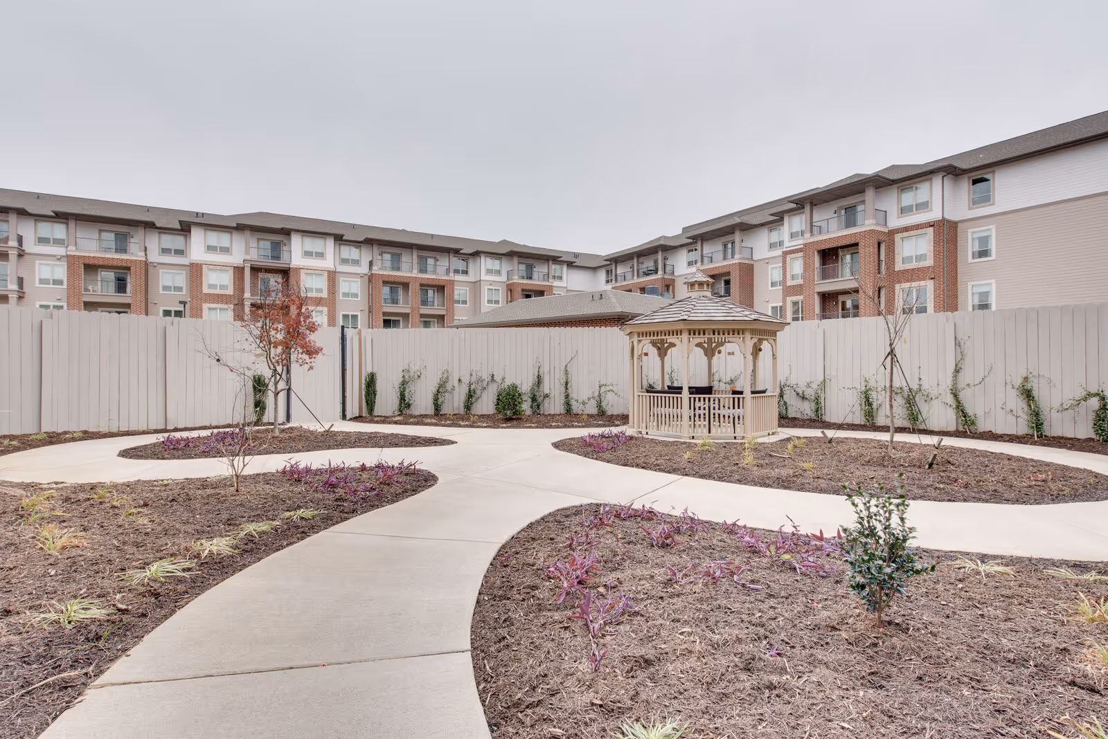Winding paved paths and landscaped beds around a wooden gazebo in a fenced courtyard with a multi-story apartment building in the background.