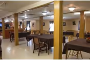 Interior dining room with multiple tables covered in dark tablecloths, wooden chairs, support columns, and a serving counter along the back wall.