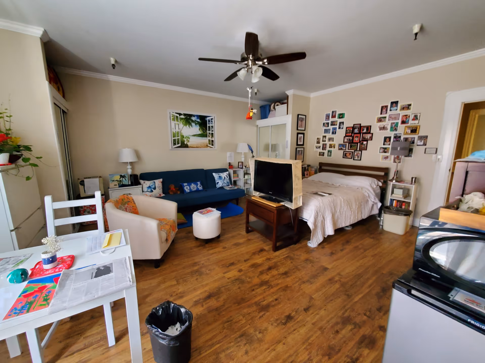 A cozy studio apartment with a bed, a blue couch, a white armchair, a small white dining table with chairs, a TV on a wooden stand, and a ceiling fan. The walls are decorated with a large photo of a beach scene and a collage of framed pictures. The floor is wooden, and there is a small kitchen area with a microwave and mini fridge visible.