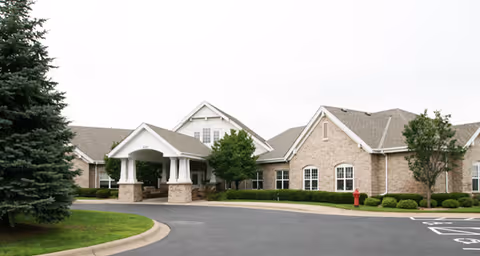 Exterior view of a single-story brick building with a covered entrance supported by white columns, surrounded by greenery and a paved driveway.