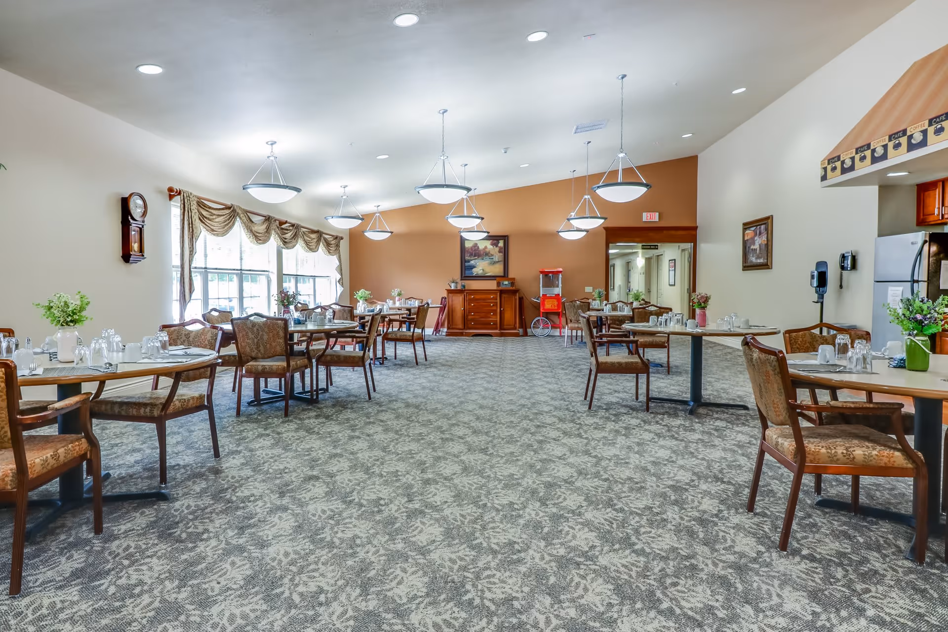 A spacious dining room in Arbor Grove Assisted Living & Memory Care with multiple tables and chairs arranged neatly. Each table is set with glasses and napkins. The room has large windows with draped curtains on the left side, several hanging light fixtures, a patterned carpet, and a warm color scheme with beige and orange walls. There is a popcorn machine and a sideboard with a framed painting on the far wall.