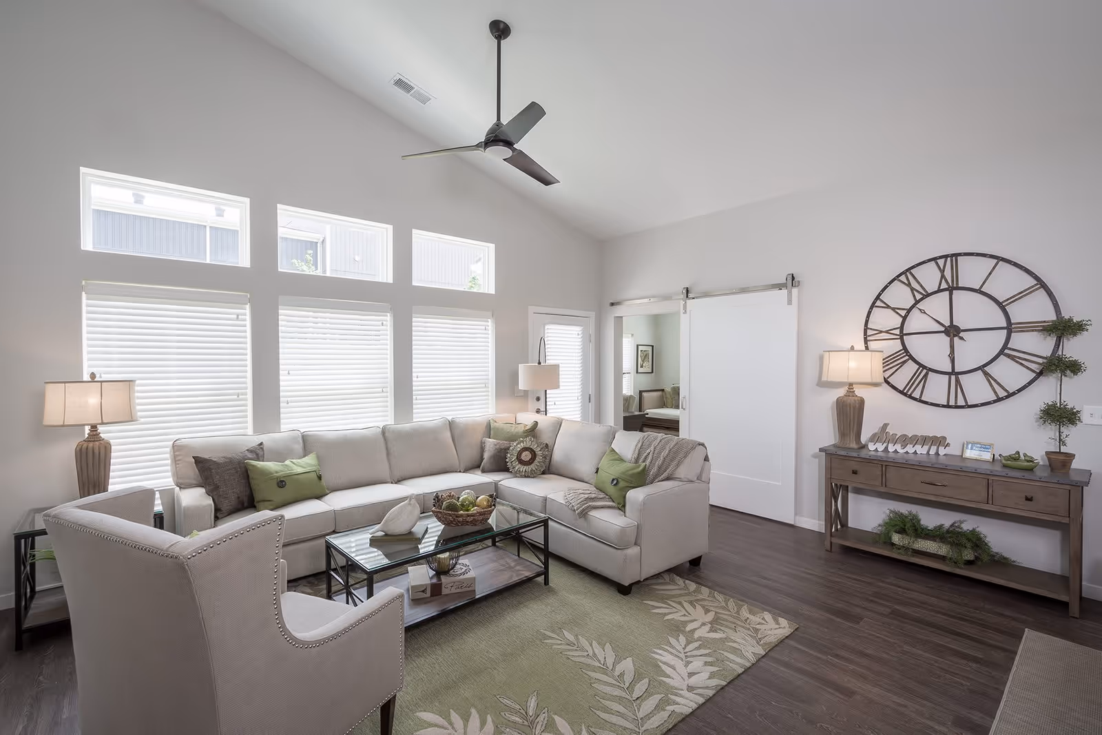 Bright and modern living room with a large beige sectional sofa adorned with green and brown pillows, a glass coffee table with decorative items, a beige armchair, a green patterned rug, a wooden console table with a large round wall clock above it, two table lamps, and a ceiling fan. The room has multiple windows with white blinds and a sliding barn door leading to another room.