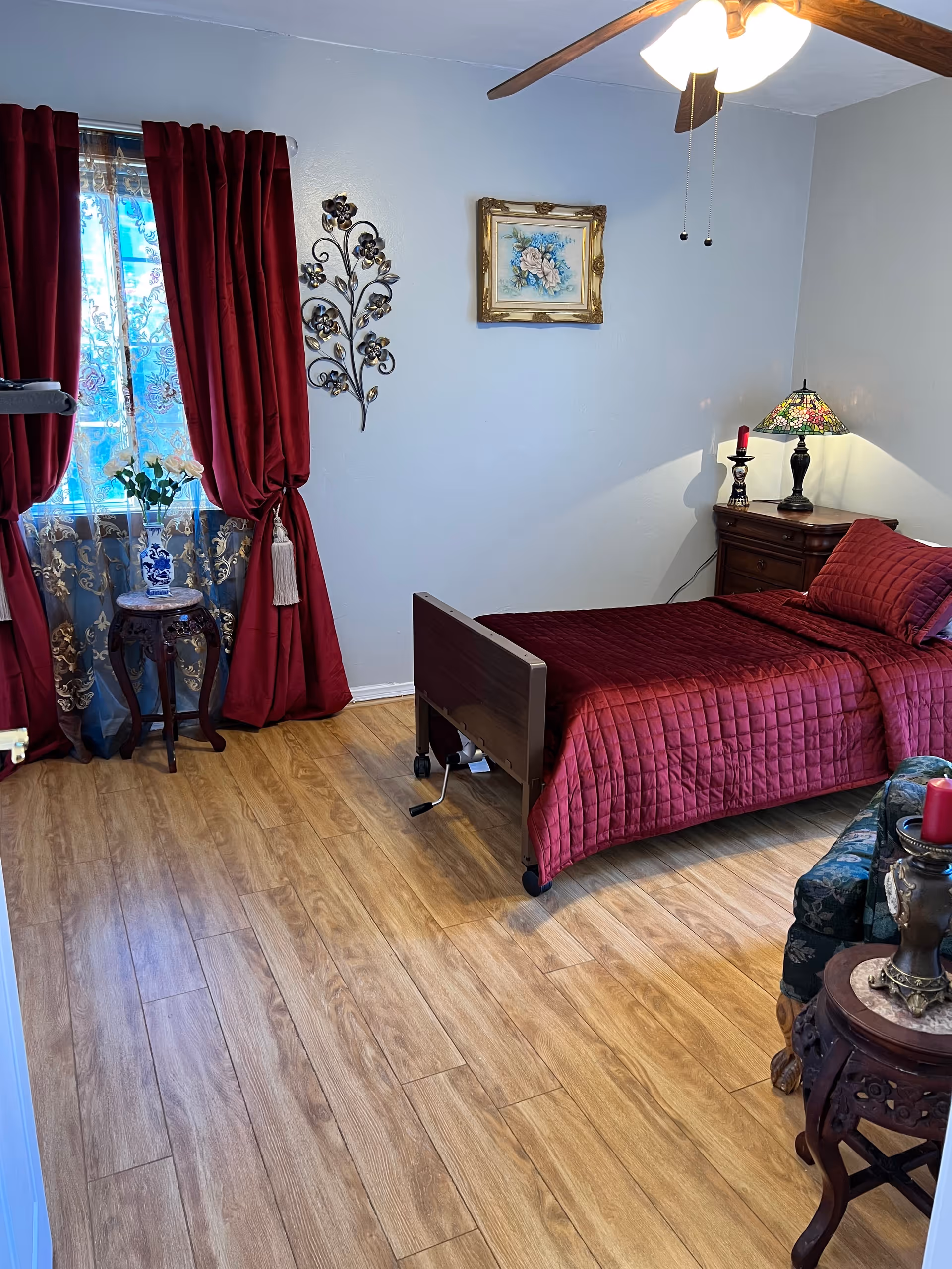 A bedroom with a single bed covered in a red quilt and matching pillow. The room has wooden flooring, a ceiling fan with lights, and a window with red curtains and sheer lace curtains underneath. There is a small wooden side table with a vase of flowers near the window, a wooden nightstand with a decorative lamp and candle, a floral armchair, and a small round wooden table with a red candle on it. A framed floral painting and a metal wall decoration hang on the light-colored wall.