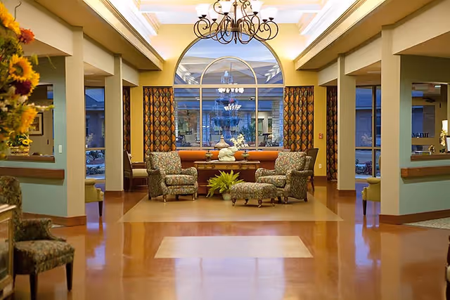 A warmly lit interior common area with polished wooden floors, featuring a seating arrangement of four patterned armchairs around a small table with decorative plants and sculptures. Large windows with patterned curtains allow natural light, and a chandelier hangs from the ceiling.