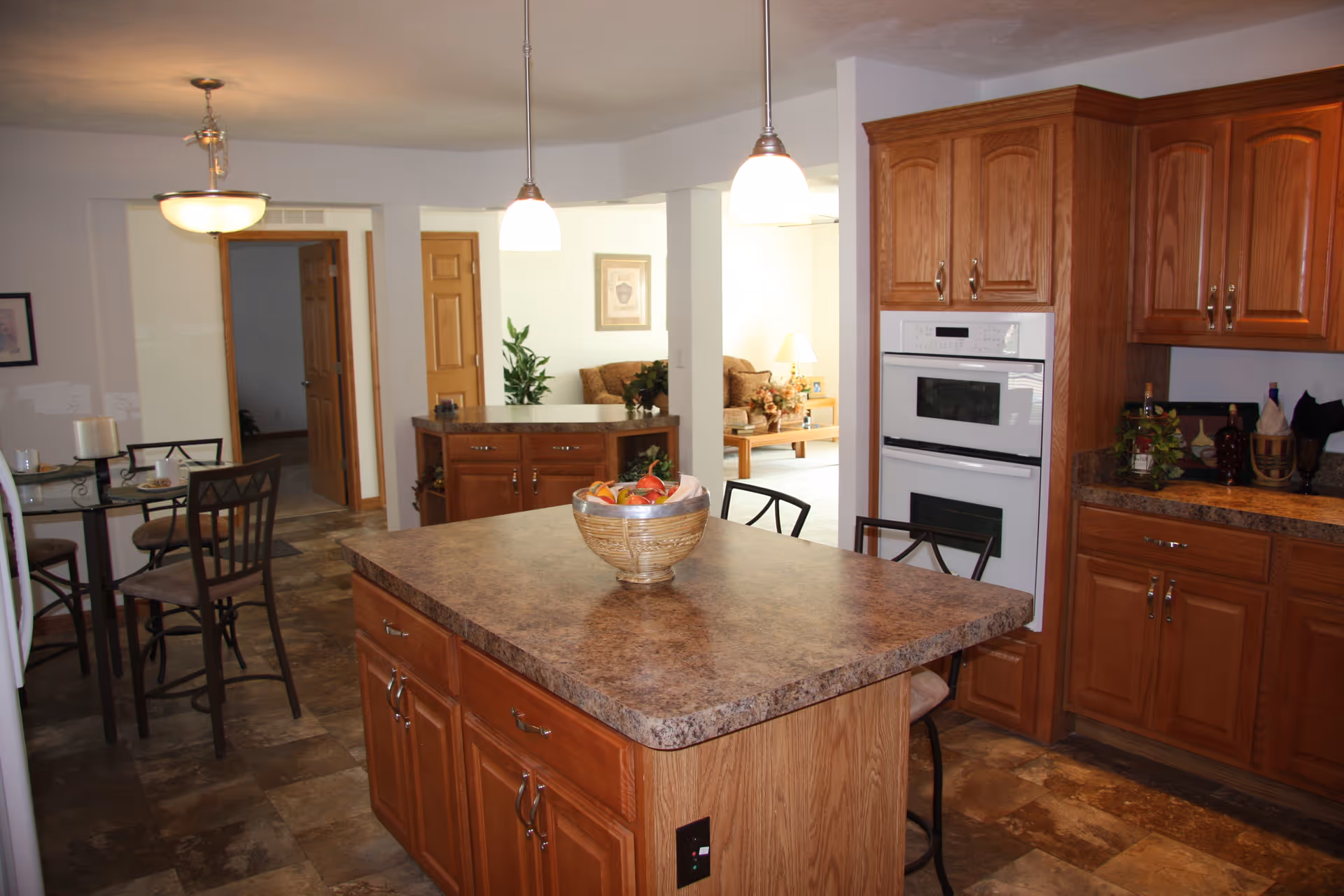 Interior view of a kitchen with a large central island topped with a basket of fruit. The kitchen features wooden cabinets, a built-in oven, and a countertop with various items. In the background, there is a small dining area with a table and chairs, and a living room area with a sofa and a coffee table.