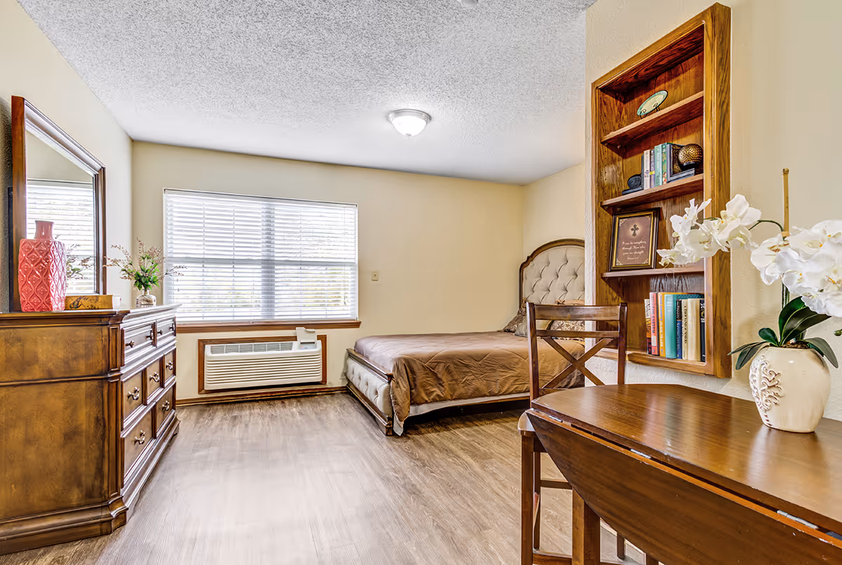 A bright bedroom with a large window covered by blinds, a bed with a tufted headboard and brown bedding, a wooden dresser with a mirror and decorative items, a wooden table with a chair, and a built-in wooden bookshelf with books and decorative pieces. The floor is wood, and the walls are painted light beige.