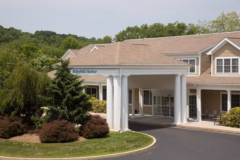 Exterior view of a senior living facility named Ridgefield Harbor, showing the entrance with a covered driveway supported by white columns, surrounded by green trees and bushes under a clear sky.