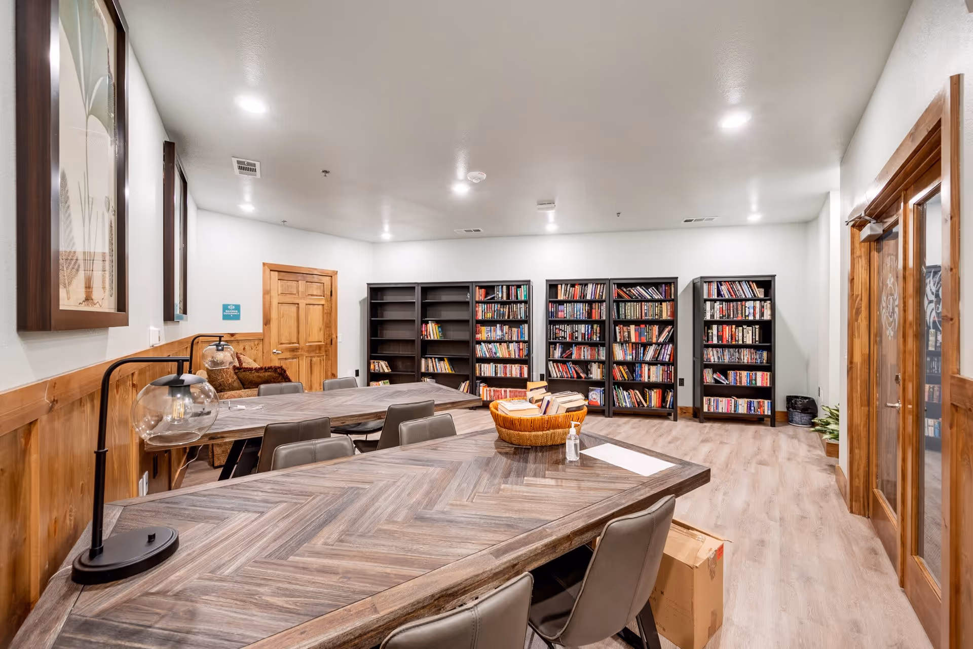 Communal library/activity room with long wooden tables and chairs facing multiple bookshelves along the back wall.