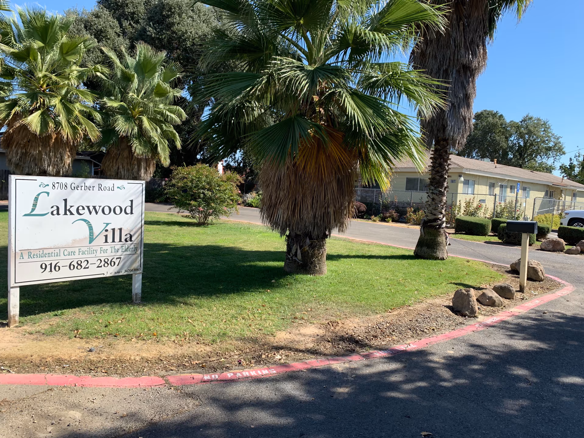 Outdoor view of Lakewood Villa, a residential care facility for the elderly, showing a sign with the facility's name, address, and phone number. The scene includes palm trees, a mailbox, a driveway, and a single-story building in the background under a clear blue sky.