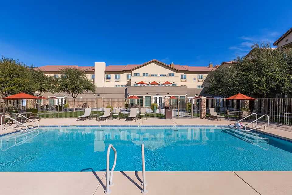 Outdoor swimming pool with clear blue water in front of a multi-story senior living facility building. Poolside lounge chairs and tables with red umbrellas are arranged around the pool. Trees and a clear blue sky are visible in the background.