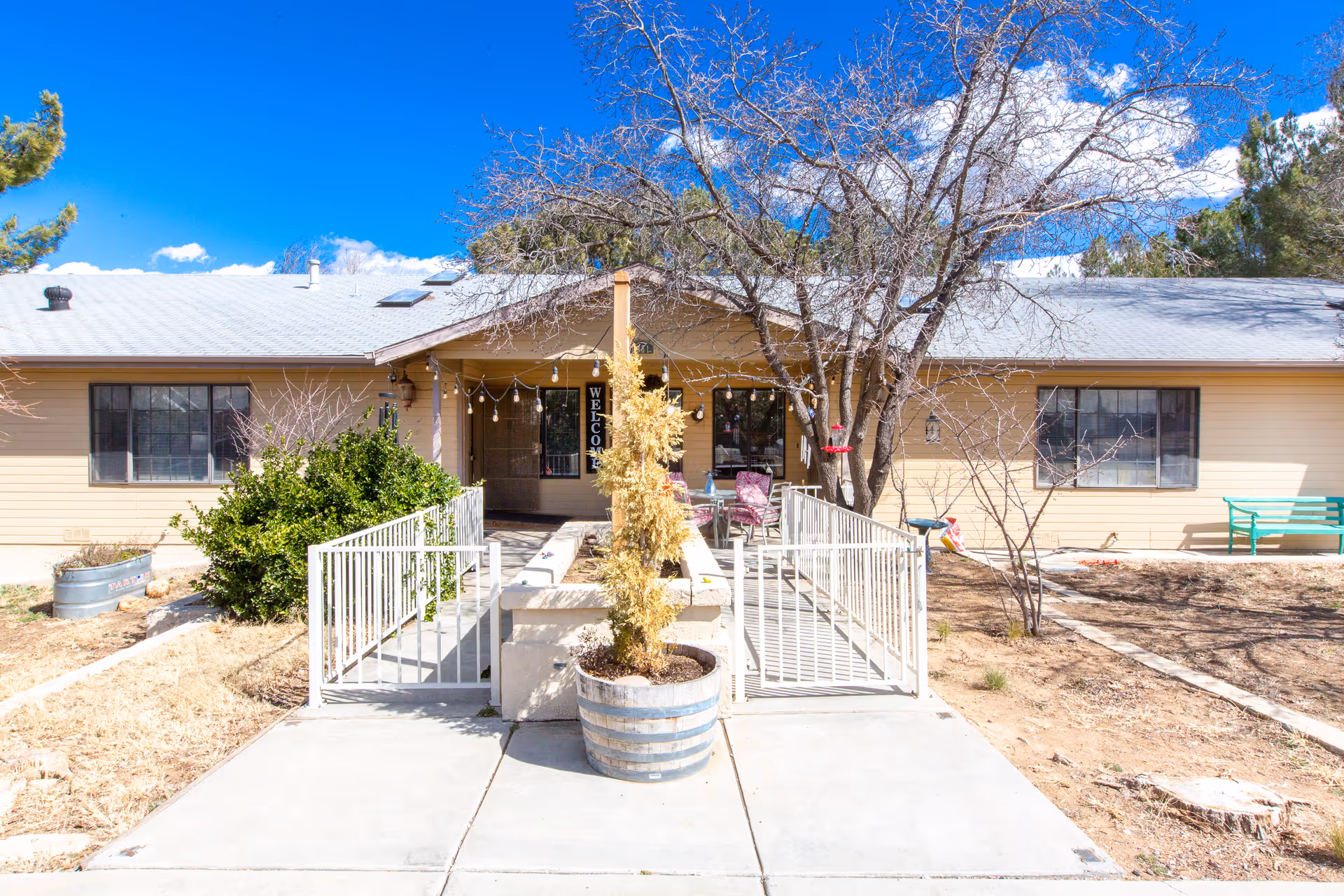 Front exterior view of a single-story assisted living facility building with a light beige exterior, a gray roof, and a wheelchair accessible ramp leading to the entrance. There are leafless trees and some bushes around the entrance area under a bright blue sky with a few clouds.