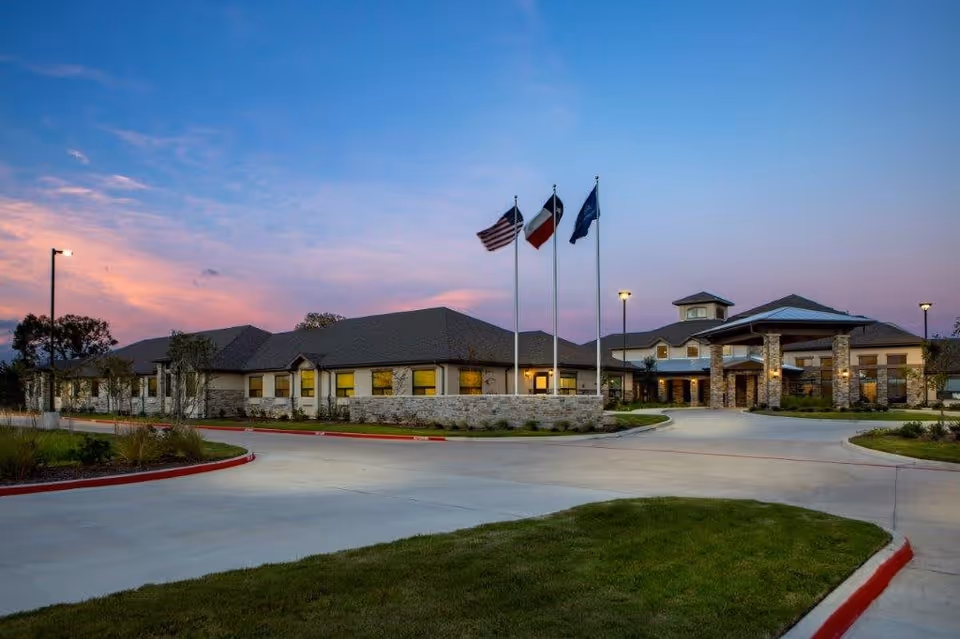 Exterior view of The Shores at Clear Lake senior living facility at dusk, showing a large building with stone and brick facade, three flagpoles with flags, well-lit entrance, and a curved driveway surrounded by landscaped grass and plants.