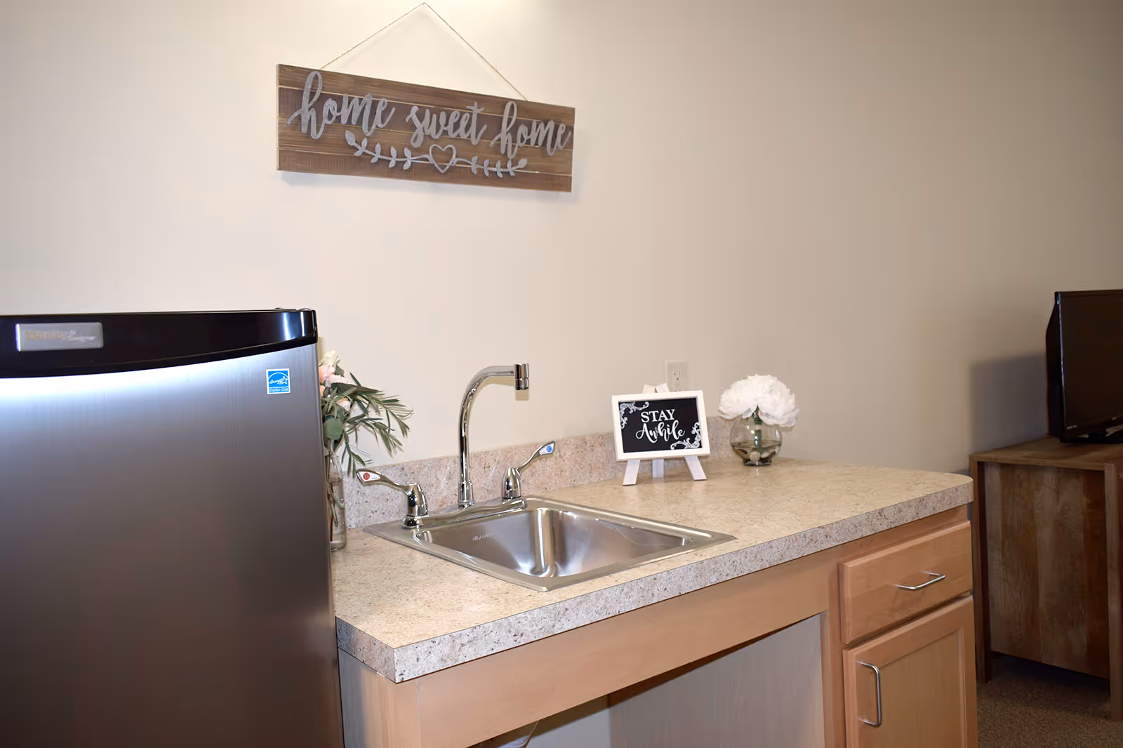 Small kitchenette with a countertop sink, mini fridge, and decorative signs including a 'home sweet home' wall plaque.