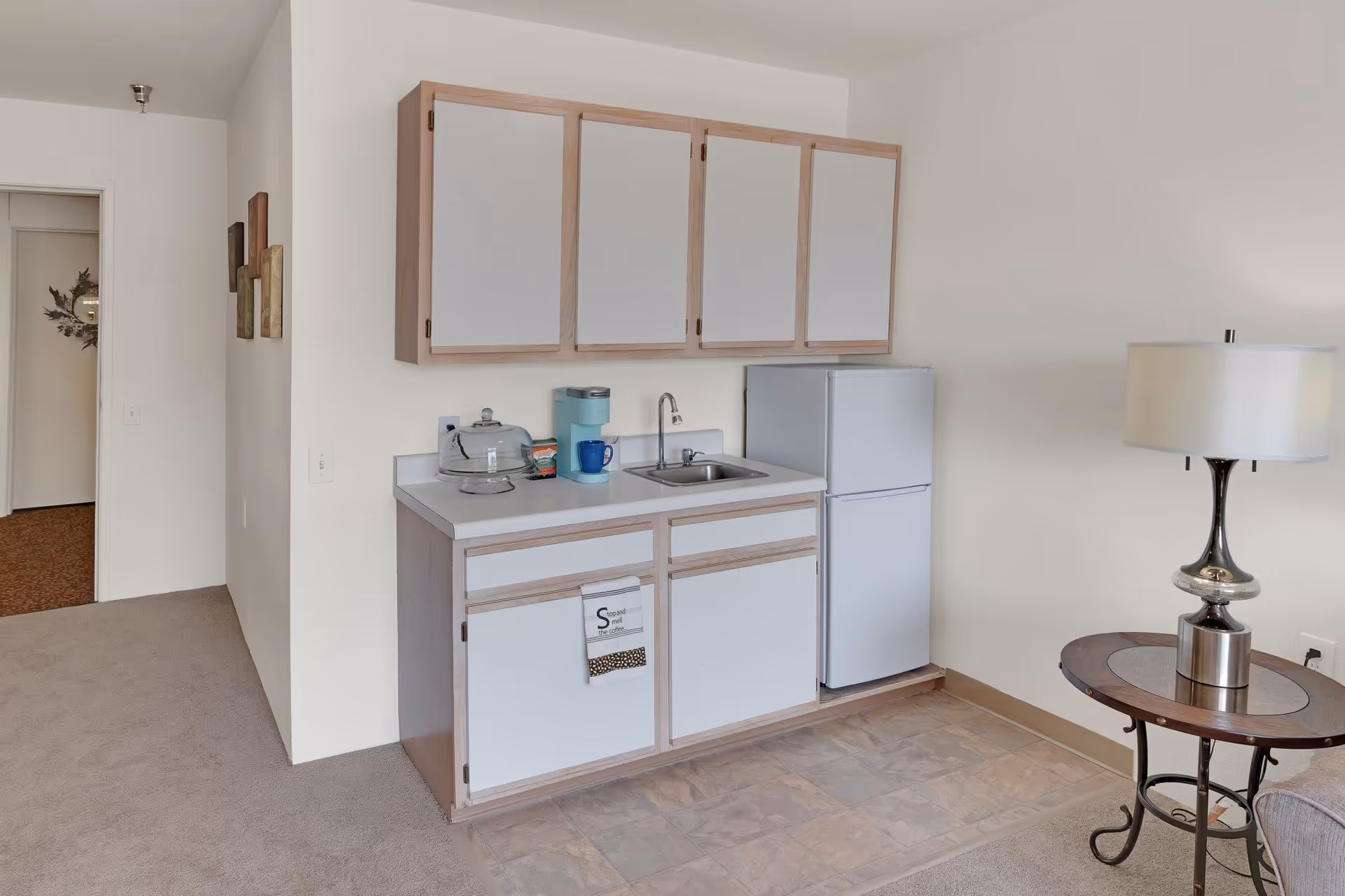 Small kitchenette area with white cabinets and a countertop. There is a small sink, a mini refrigerator, a coffee maker, a glass cake stand with a cover, and a blue mug on the counter. To the right, there is a round side table with a silver lamp on it. The floor is a mix of carpet and tile, and the walls are painted white.