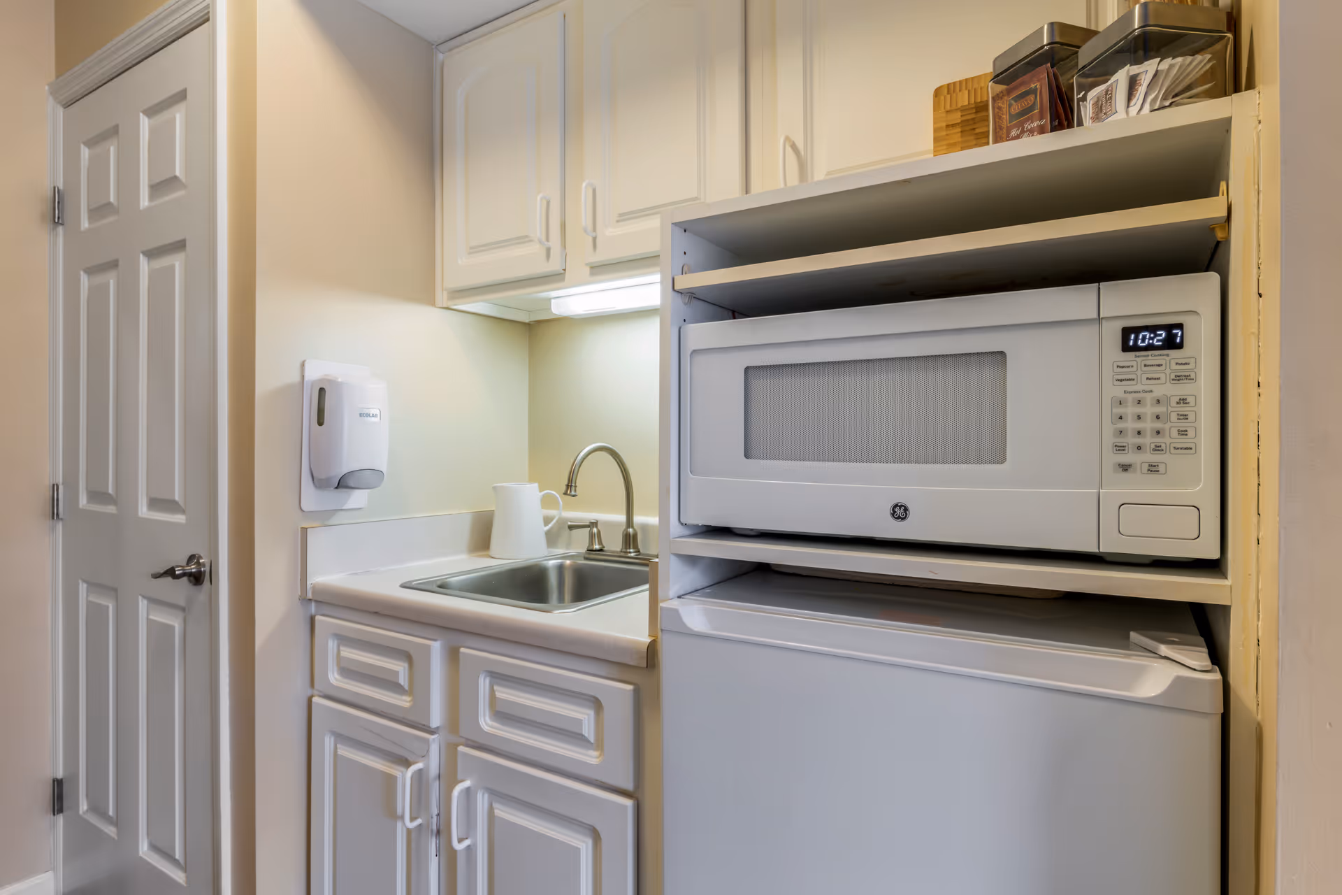 Small kitchenette area with white cabinets, a stainless steel sink with a faucet, a white microwave oven placed above a white mini refrigerator, and a white door to the left. There is a soap dispenser mounted on the wall above the counter and some containers on the shelf above the microwave.