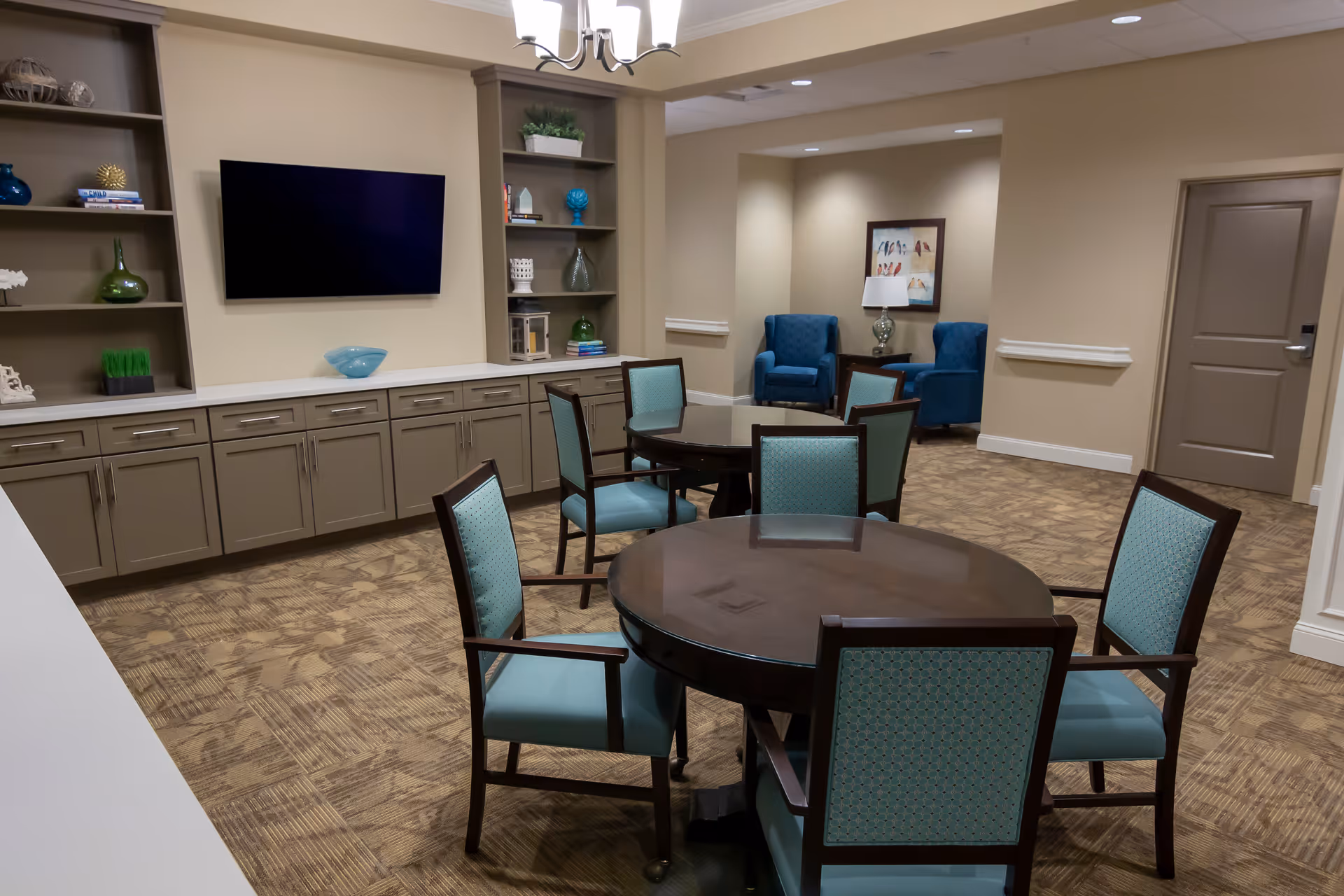 A common area in a senior living facility featuring round tables with teal upholstered chairs, built-in cabinets and shelves with decorative items, a wall-mounted flat screen TV, and a seating nook with two blue armchairs and a side table with a lamp.