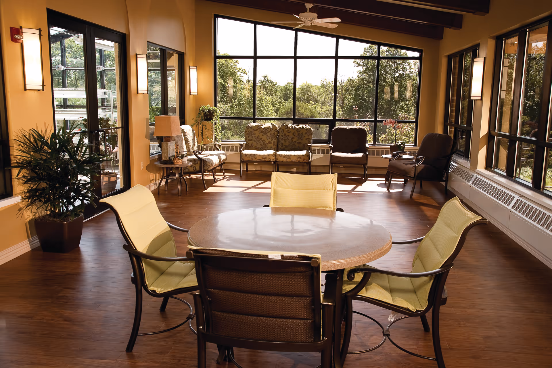 Sunlit communal lounge with a round table and chairs in the foreground and cushioned seating along large windows overlooking trees.