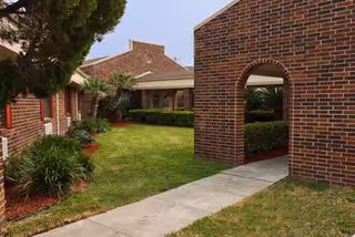 Brick buildings surround a sunny outdoor courtyard with a concrete path and shrubs.