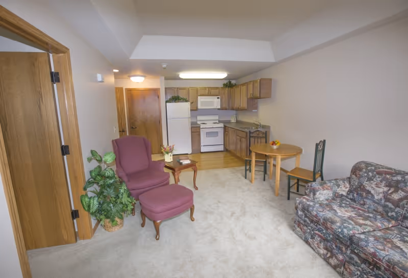 Interior view of a senior living apartment featuring a small kitchen with wooden cabinets, a white refrigerator, stove, and microwave. The living area includes a floral patterned sofa, a purple armchair with matching ottoman, a small wooden side table with a flower vase and books, a round wooden dining table with two chairs, and a potted plant near an open wooden door.