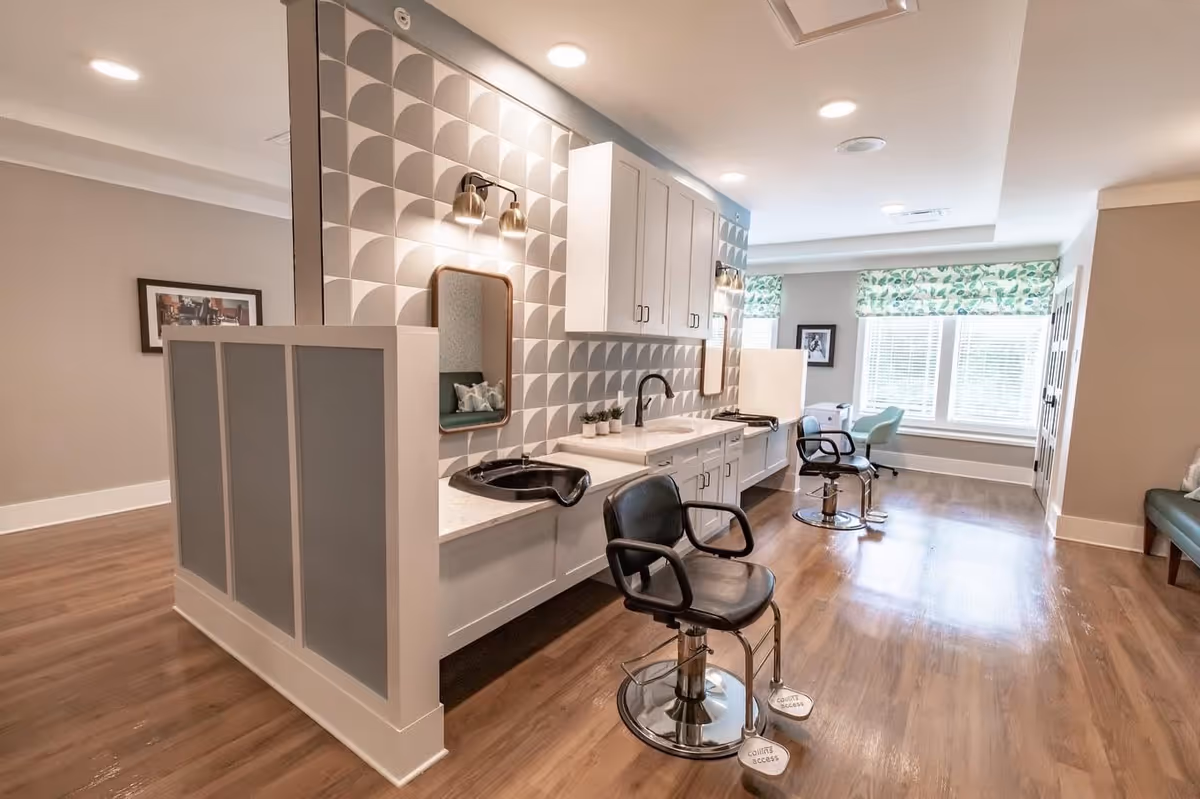 Interior view of a salon area in a senior living facility with two black salon chairs in front of sinks and mirrors mounted on a wall with a geometric pattern. The room has wooden flooring, white cabinetry, and large windows with green patterned valances allowing natural light to enter.