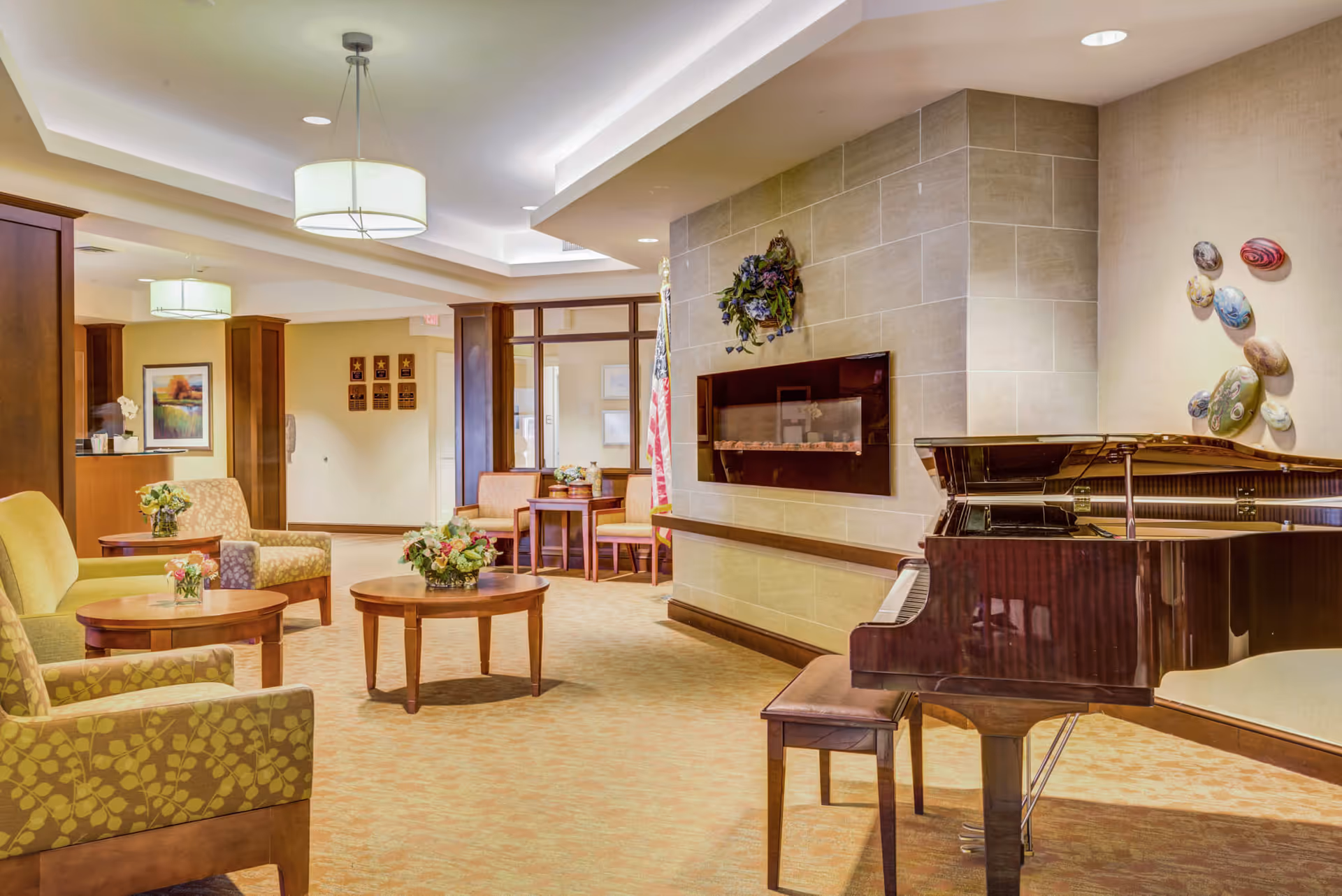 A cozy senior living common area with a grand piano on the right, a modern electric fireplace mounted on a tiled wall, and comfortable seating including armchairs and small round tables with flower arrangements. The room has warm lighting and decorative wall art, creating a welcoming atmosphere.