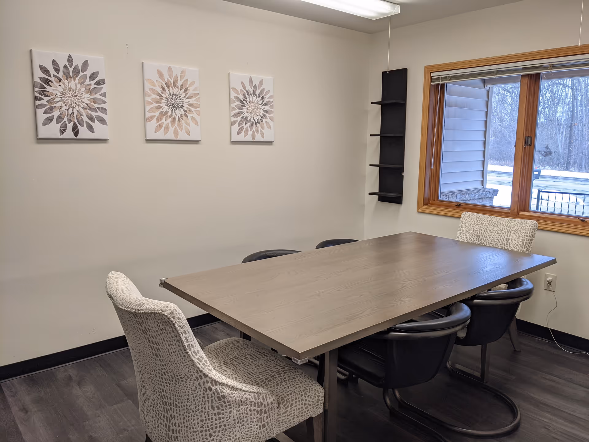 A small meeting or dining room with a rectangular wooden table surrounded by six chairs, two upholstered with a light patterned fabric and four black chairs. The room has light-colored walls with three floral artwork pieces hanging on one wall. There is a window with a wooden frame showing an outdoor view of trees and snow. A black wall-mounted shelf is in the corner.