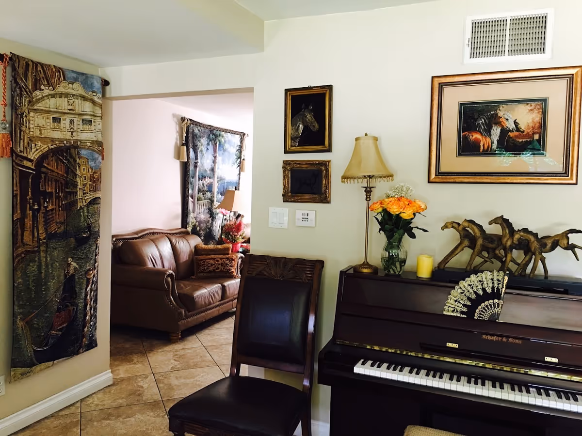 A cozy living room area featuring a brown leather sofa, a dark wooden chair, and a piano with decorative horse figurines and a vase of orange roses on top. The walls are adorned with framed horse artwork and a standing lamp. Tapestries with scenic designs hang on the walls.