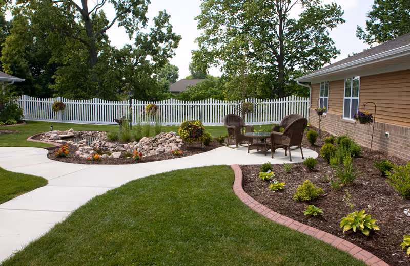 Outdoor garden area with a white picket fence, green grass, a small pond surrounded by rocks, flower beds, and a patio with wicker chairs and a glass-top table next to a building with tan siding and brick foundation.