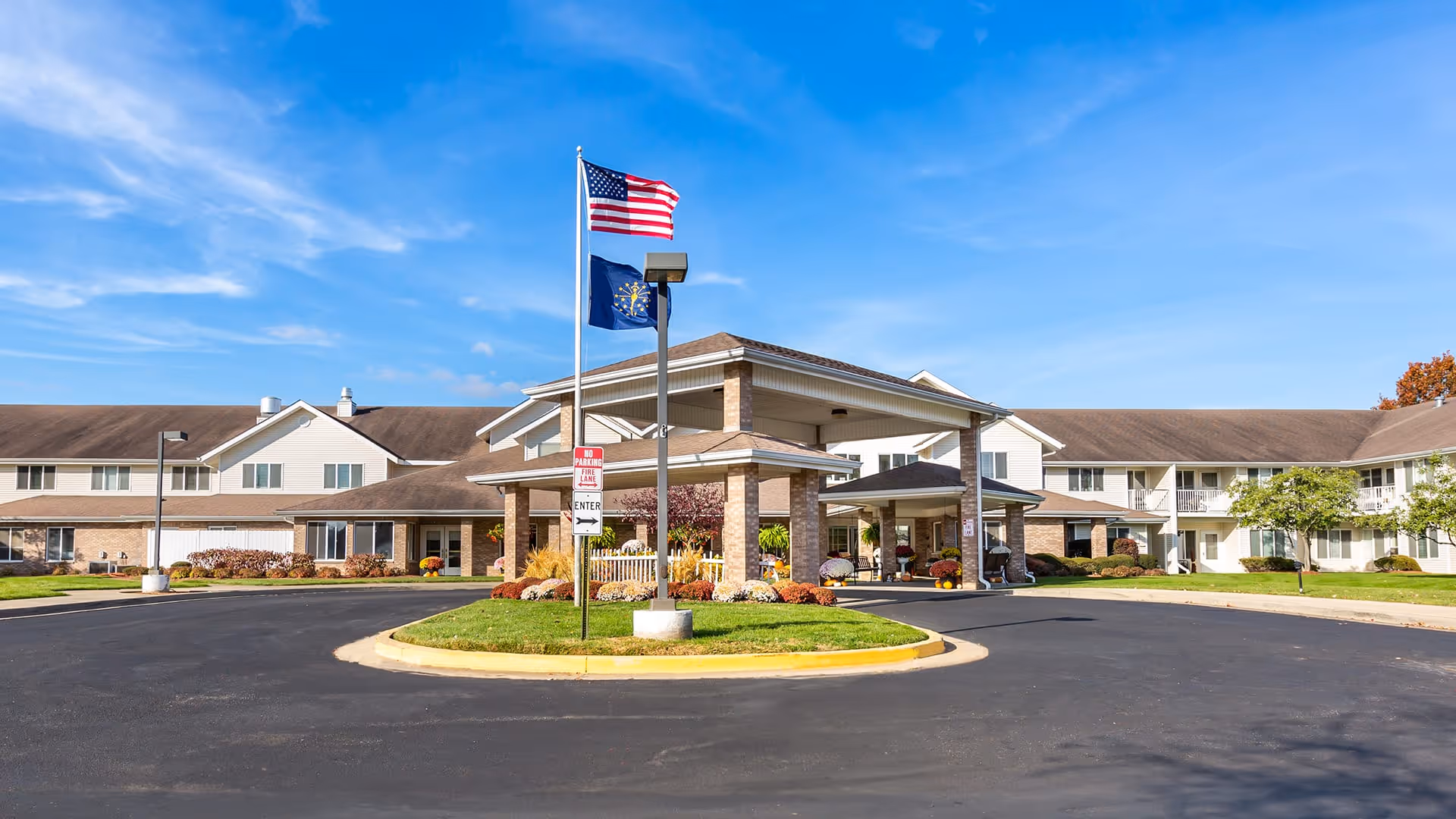 Front exterior view of Independence Village of Columbus, showing a large building with a covered entrance, two flagpoles with the American and Indiana state flags, a circular driveway, and well-maintained landscaping under a clear blue sky.