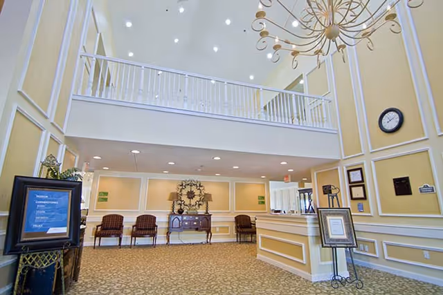 Interior view of a senior living facility lobby with a high ceiling and a chandelier. The room features a reception desk on the right, a decorative console table with lamps and a wall clock on the back wall, and several chairs lined up against the wall. The walls are painted yellow with white trim, and there is a balcony railing visible above.