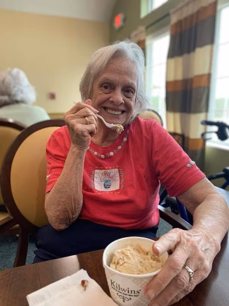 An elderly woman with white hair wearing a red shirt is sitting at a table in a dining area, smiling and holding a spoon with ice cream near her mouth. She has a cup of Kilwins ice cream in front of her. The background shows another person seated and windows with striped curtains.