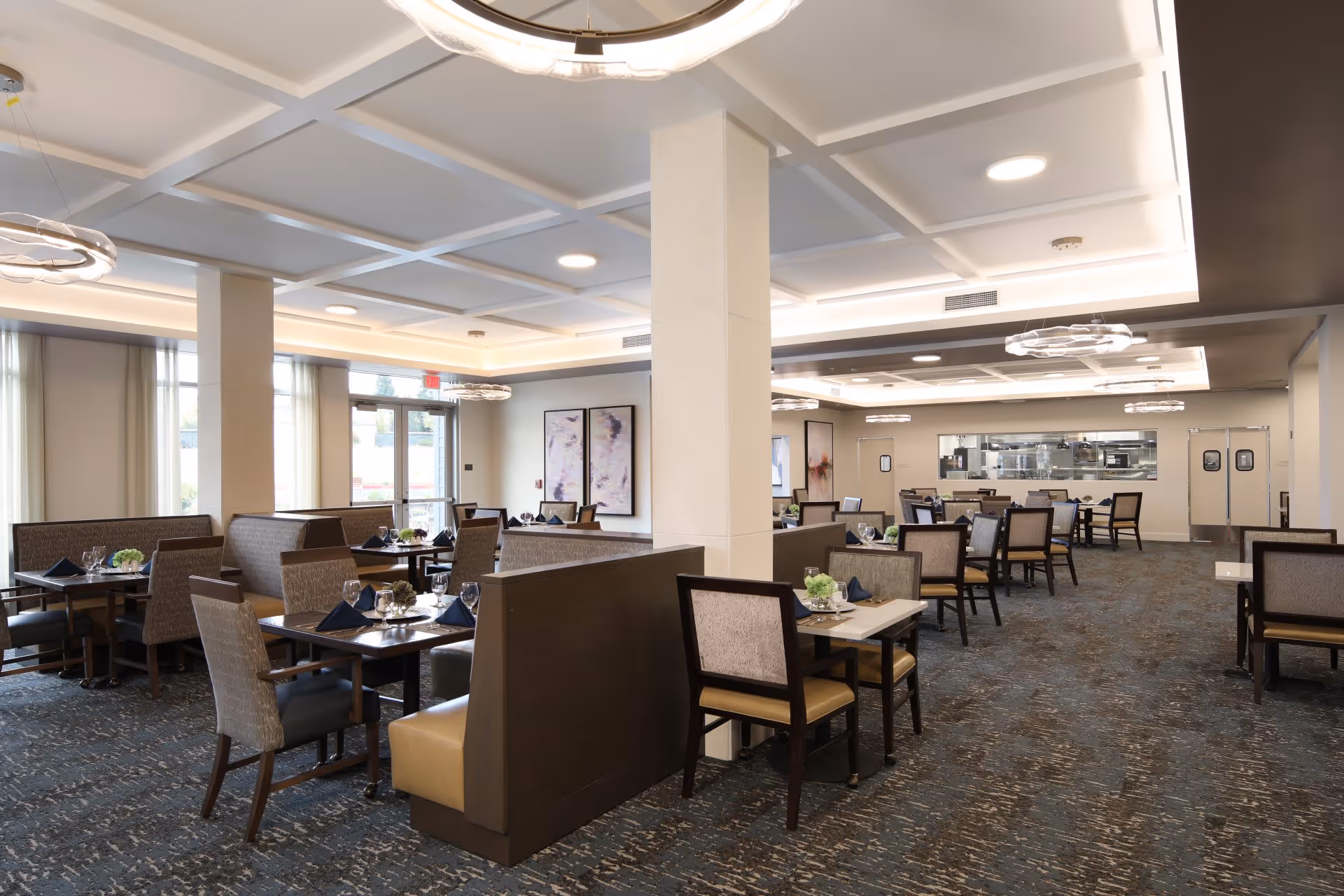A spacious and well-lit dining room in an assisted living facility with multiple tables and chairs arranged neatly. The tables are set with glasses, napkins, and small floral centerpieces. The room features modern ceiling lights, large windows with curtains, and artwork on the walls. There is a visible kitchen area in the background with double doors.