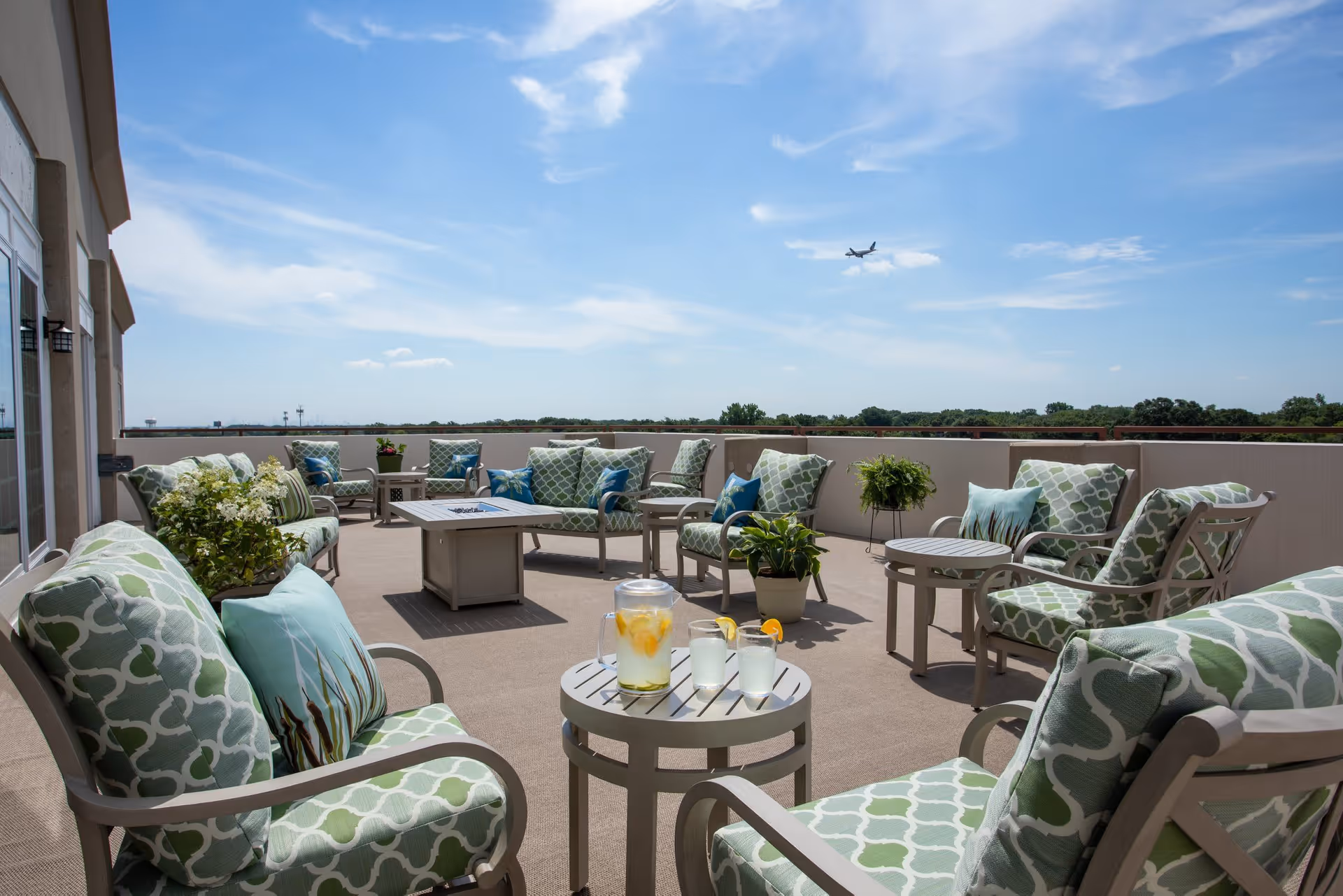 Outdoor patio area with cushioned chairs and small tables arranged in a circle under a clear blue sky. There are potted plants and a pitcher of lemonade with glasses on one of the tables. An airplane is visible flying in the sky in the background.