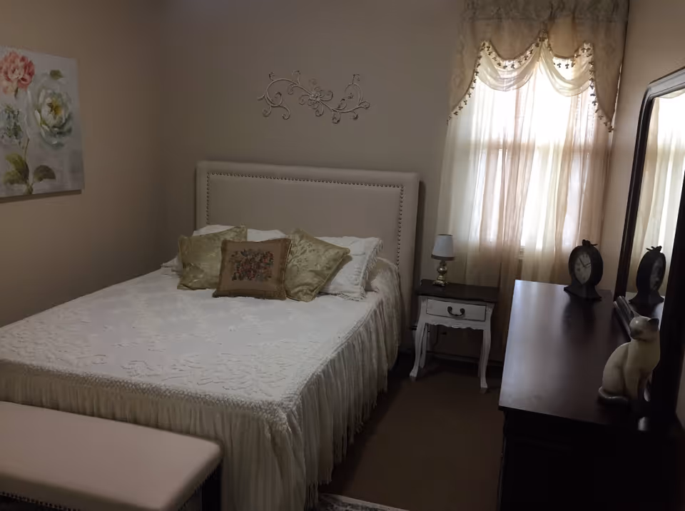 Cozy bedroom with a made bed, decorative pillows, nightstand, dresser with mirror, and a curtained window.