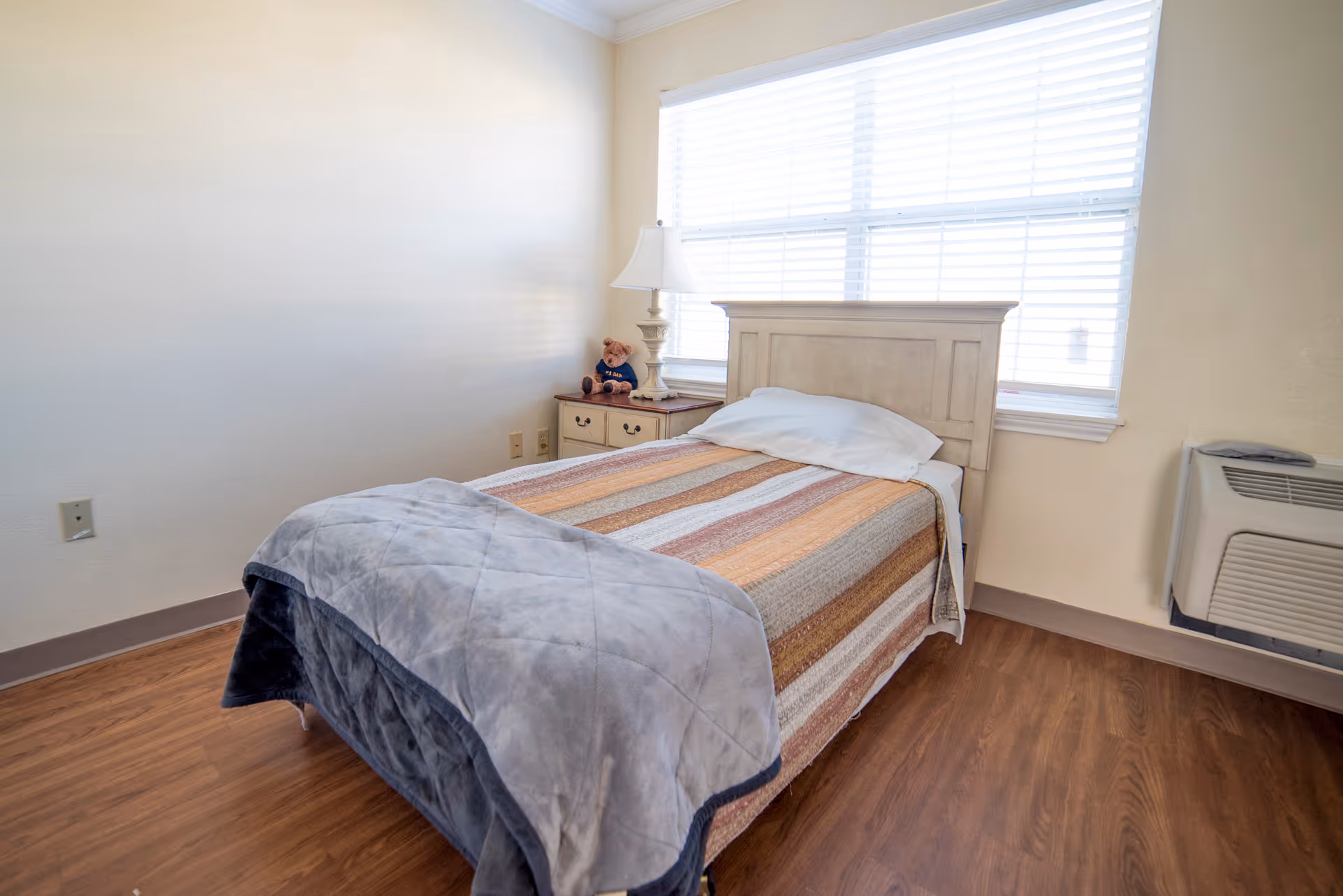 Small bedroom with a single bed covered in a striped bedspread, a nightstand with a lamp and teddy bear, a large window with blinds, and a wall air unit.