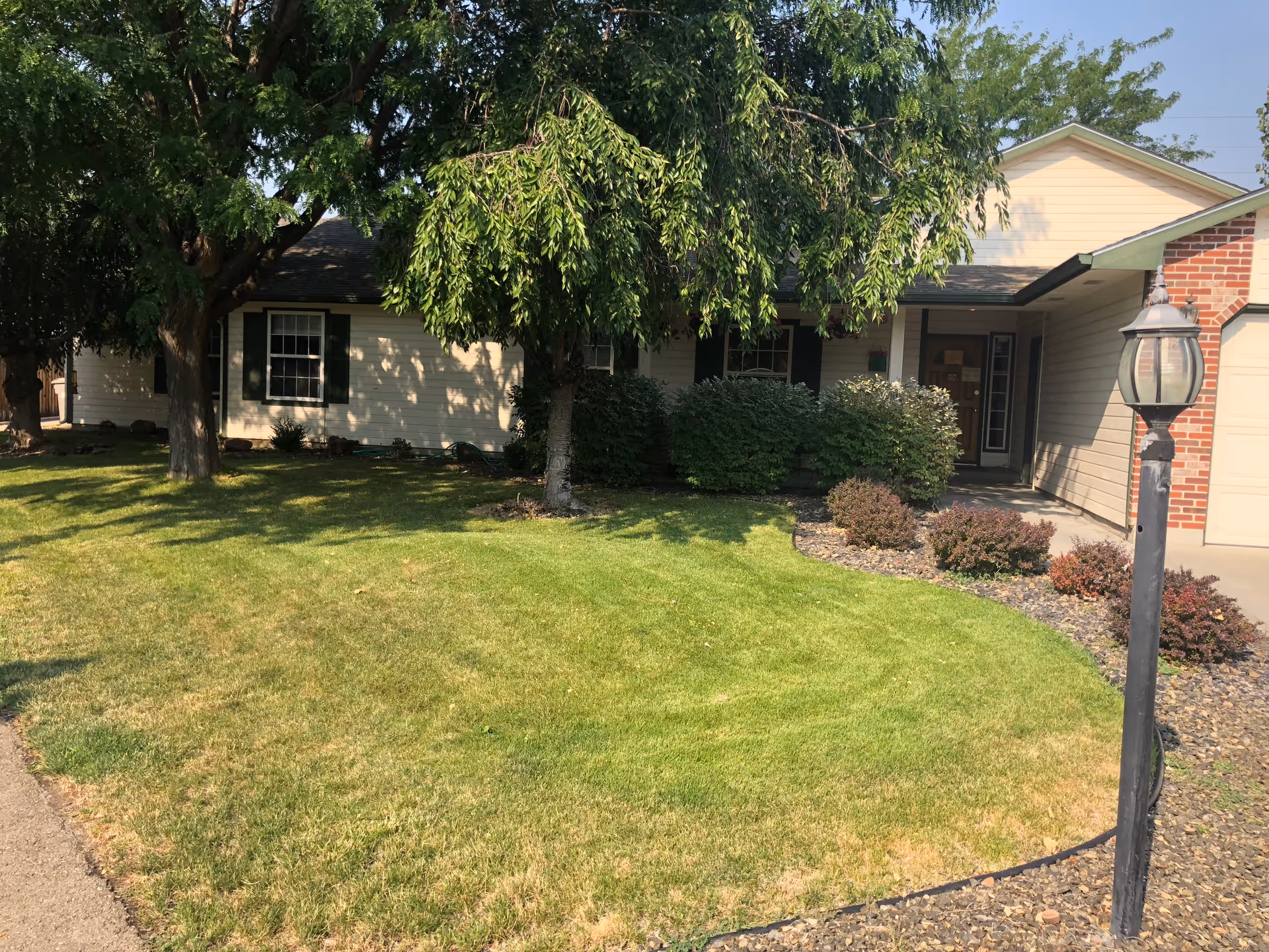 Front exterior view of a single-story house with beige siding, green shutters, and a brick accent near the garage. The house is surrounded by a well-maintained lawn, trees, bushes, and a black lamp post near the driveway.