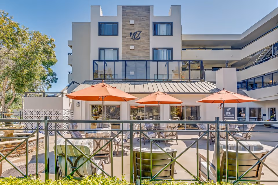 Outdoor patio area at Burlingame Senior Living with several tables and chairs under orange umbrellas, surrounded by a metal railing. The building in the background has multiple floors with balconies and large windows, and a sign indicating the main entrance.