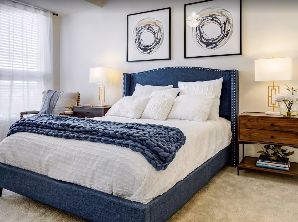 A neatly made bed with a blue upholstered headboard and white bedding, adorned with multiple white pillows and a chunky blue knit throw blanket. On either side of the bed are wooden nightstands with modern lamps. A cushioned chair with a gray throw is placed near a window with sheer white curtains. Two abstract circular artworks hang above the bed.