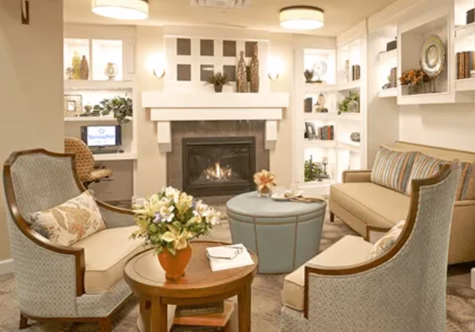 Cozy living room with a lit fireplace, two upholstered armchairs, a sofa, round ottoman, coffee table with flowers, and built-in shelving.
