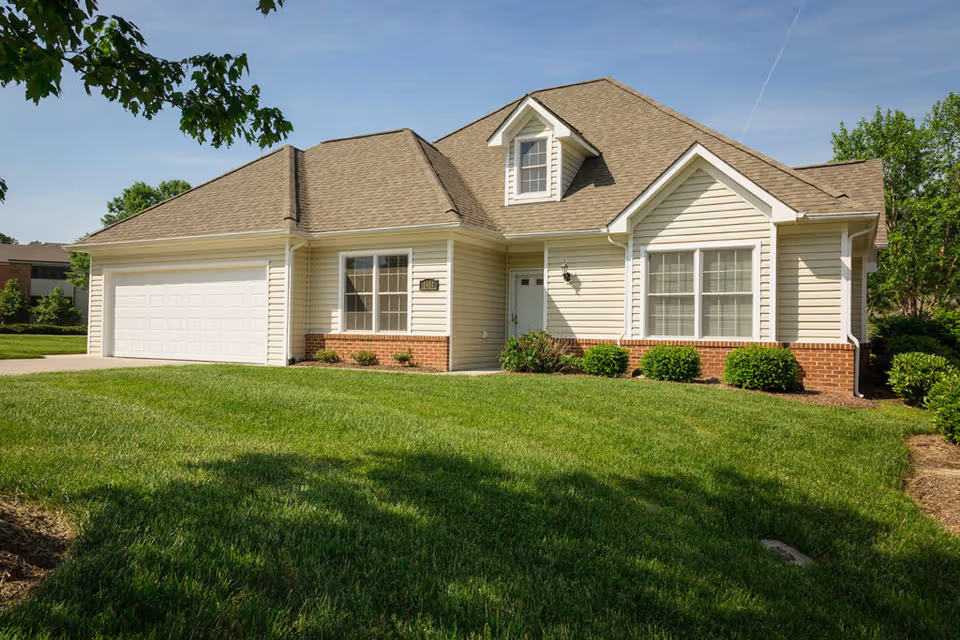 Single-story house with beige siding and brick accents, a two-car garage, and a well-maintained green lawn under a clear blue sky.