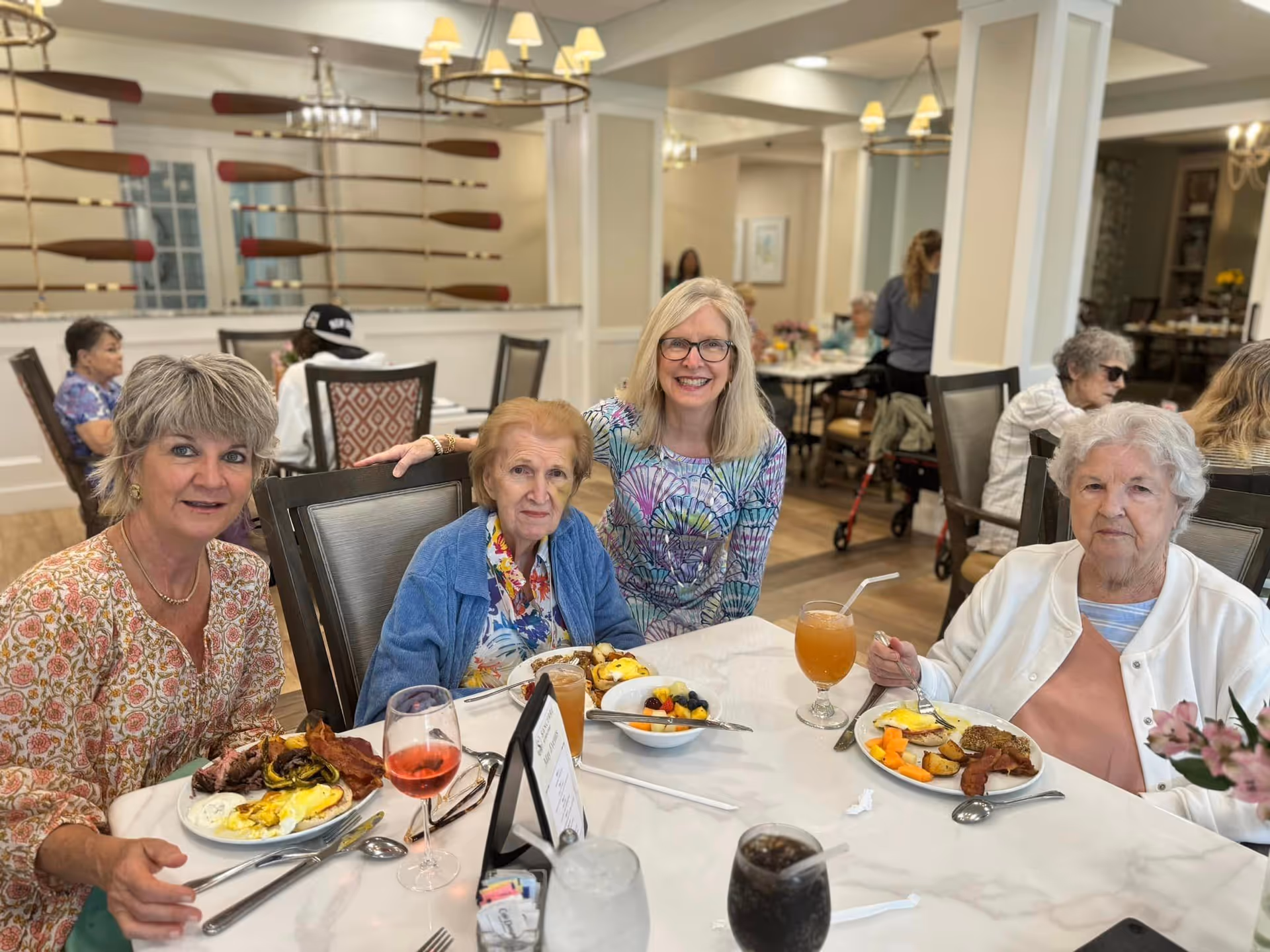 Four women sitting around a dining table in a senior living facility, enjoying a meal together. The table is set with plates of food including eggs, bacon, fruit, and beverages. The room has a warm, inviting atmosphere with decorative paddles on the wall and other residents dining in the background.