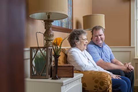 An elderly woman and a middle-aged man sitting together on a patterned sofa in a warmly lit living room. There are two table lamps with beige shades, a small wooden box, a vase with orange flowers, and a framed picture on the wall behind them.