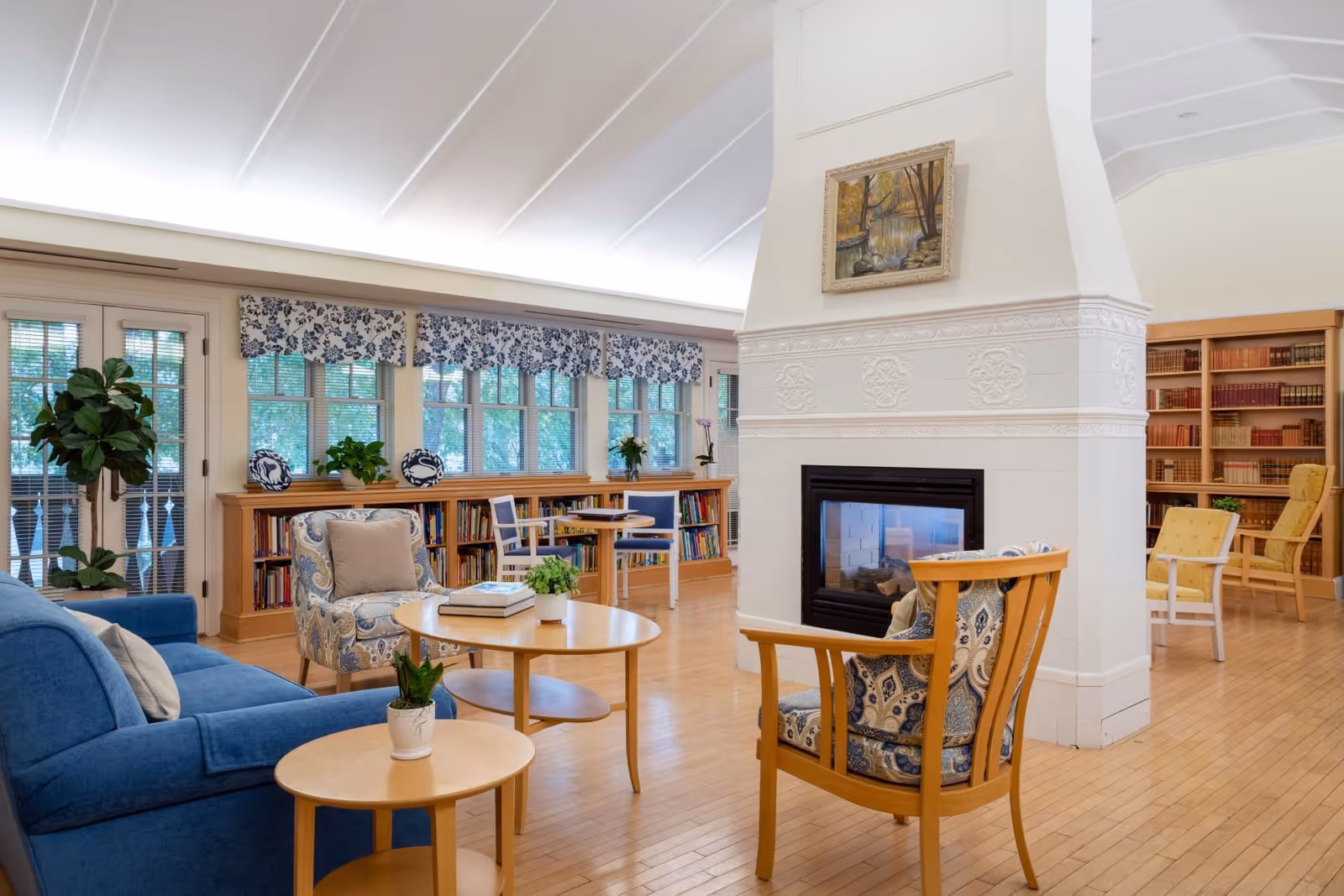 A cozy living room area in the Scandinavian Living Center featuring a blue sofa, patterned armchairs, wooden tables with potted plants, a white fireplace with a painting above it, bookshelves filled with books, and large windows with floral valances letting in natural light.