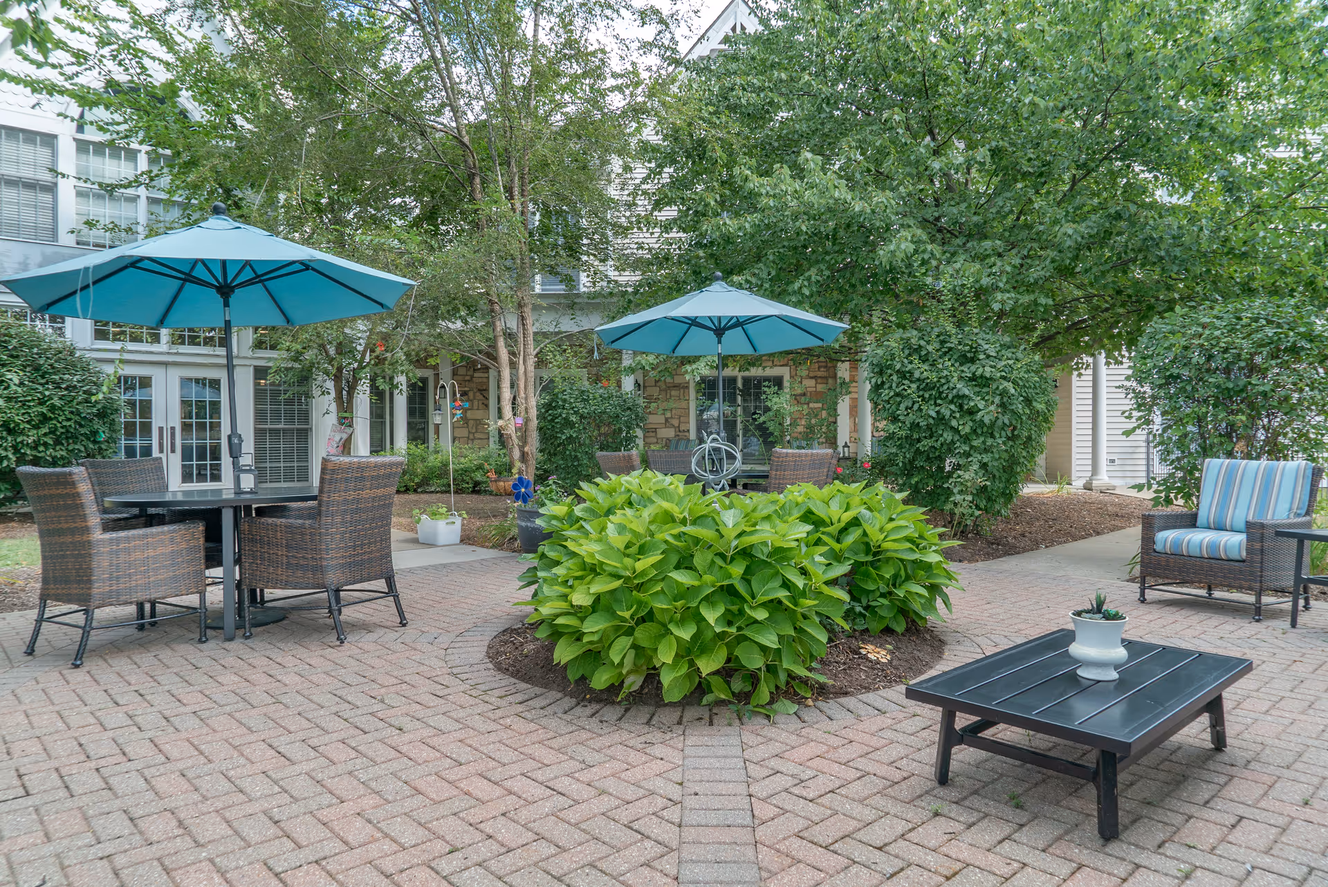 Outdoor patio area with brick paving, two tables with blue umbrellas and wicker chairs, surrounded by green bushes and trees. A low black table with a small potted plant and a wicker chair with blue and white striped cushions are also visible. The building with windows and doors is in the background.