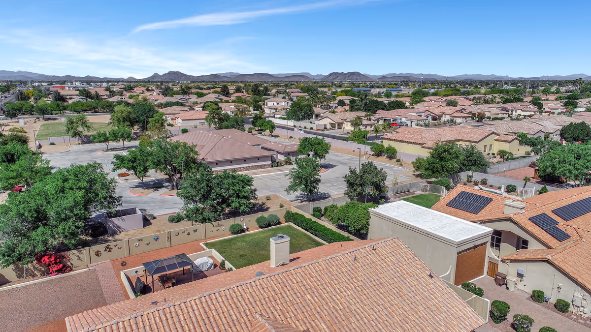 Aerial view of a residential neighborhood with multiple houses featuring tiled roofs, green trees, and a parking lot. The landscape includes distant mountains under a clear blue sky.
