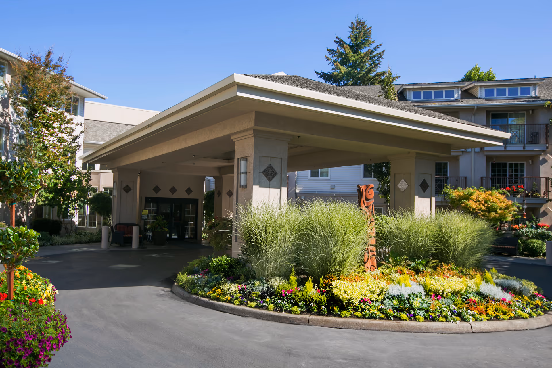 Entrance to The Lakeshore senior living facility with a covered driveway, surrounded by well-maintained landscaping including colorful flowers, ornamental grasses, and shrubs. The building exterior is visible with balconies and windows under a clear blue sky.