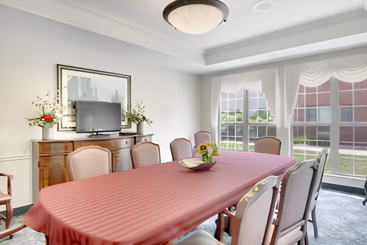 A dining room with a long table covered by a red tablecloth surrounded by cushioned chairs. There is a wooden sideboard with a TV and floral arrangements on top. Large windows with white curtains allow natural light to fill the room.